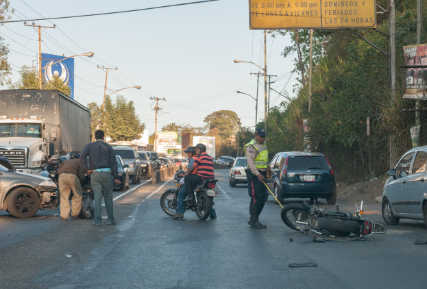 Eine Gruppe von Menschen steht in der Nähe eines verunglückten Motorrads am Straßenrand mit mehreren Fahrzeugen, darunter ein Lastwagen, im Hintergrund und Bäumen, Pfosten, Laternen, Schildern und dem Himmel.