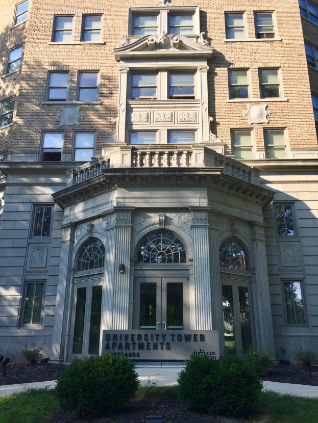Das University Tower Apartments in Chicago, Illinois, ein großes Gebäude mit Glasfenstern und -türen, umgeben von saftigem Grün und Pflanzen, mit einer Tafel mit Text davor und dem Himmel im Hintergrund.