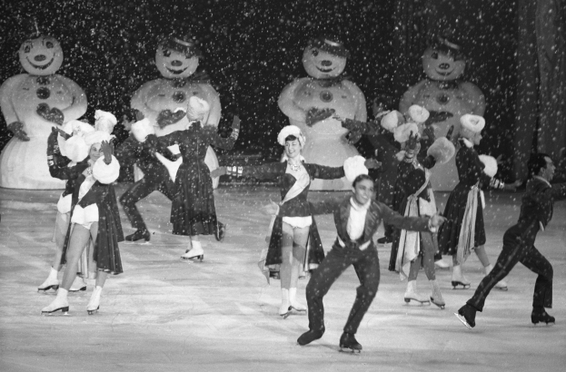 Eine Gruppe von Menschen in Kostümen und Eislaufschuhen fährt auf einer Eisbahn im Schnee, mit Schneemännern im Hintergrund, in einem Schwarz-Weiß-Bild.