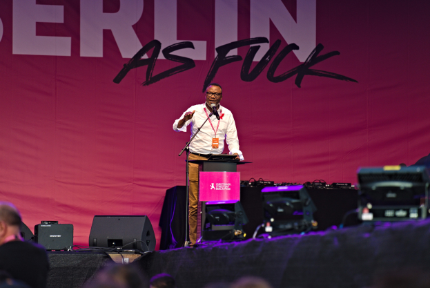 Ein Mann in weißem Hemd und braunen Hosen steht auf einer Bühne und hält ein Mikrofon, hinter ihm eine pinkfarbene Fahne mit der Aufschrift "Berlin as Fuck" und Publikum vorne.