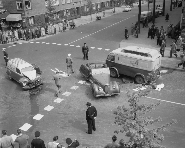 Schwarz-weiß-Szene eines Autounfalls an einer Straßenkreuzung mit mehreren Fahrzeugen, darunter ein Van, und einer Gruppe von Schaulustigen; Gebäude, Bäume, Pfosten und Texttafeln sind im Hintergrund zu sehen.