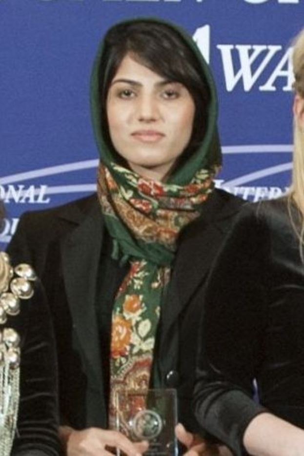 Malala Yousafzai, eine Nobelpreisträgerin für den Frieden, steht in der Mitte in einem schwarzen Kleid und einem Tuch, hält eine Auszeichnung und wird von zwei Frauen in schwarzen Kleidern flankiert, mit einem blauen Banner im Hintergrund.