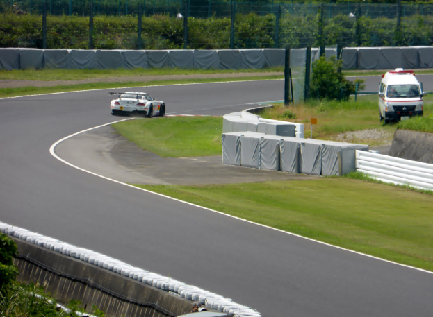 Ein Rennwagen fährt auf einer Rennstrecke mit einem Polizeiwagen im Hintergrund, umgeben von Grünflächen und Absperrungen, mit Text am unteren Bildrand.