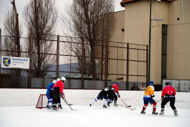 Menschen beim Eis-hockey auf einem Eisstadion mit Gebäuden, Bäumen, einer Straßenlaterne, einem Namensschild und Zäunen im Hintergrund unter einem Himmel.