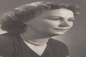 Ein Schwarz-Weiß-Foto einer Frau mit ernster Miene, die eine Kette und ein einfaches Kleid mit hohem Kragen und langen Ärmeln trägt, ihr Haar ist zurückgebunden und ihre Augen sind konzentriert.