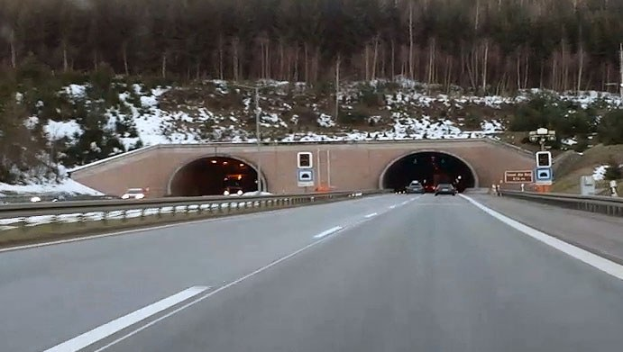 Autobahn mit Autos, die durch einen Tunnel fahren, flankiert von Geländern und Schildern, Schnee auf dem Boden, Bäume und Masten im Hintergrund und sichtbarer Himmel darüber.