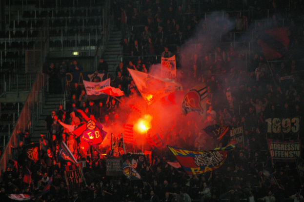 Eine große Menschenmenge in einem Stadion mit Fahnen, Bannern und einem zentralen Feuer, beleuchtet von Stadionlampen.