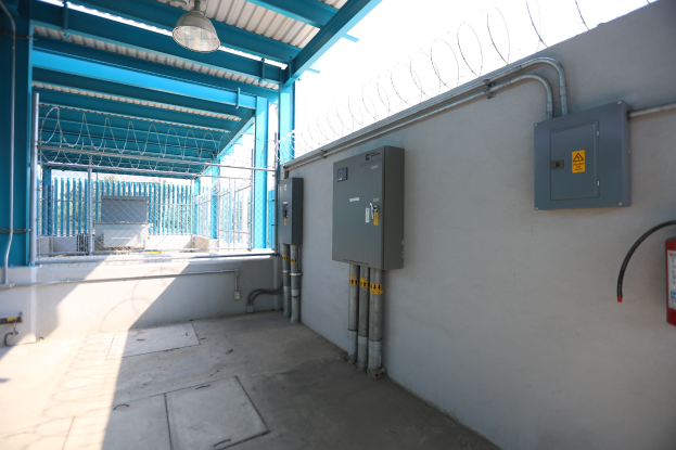 Ein Raum mit einem Feuerlöscher auf der rechten Seite, elektrischen Schächten und Rohren an der Wand, einer Decke mit einer Lampe öber und einem Zaun im Hintergrund, ähnlich einer elektrischen Umspannstation.