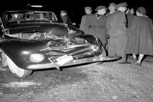 Ein Schwarz-Weiß-Bild eines beschädigten Autos am Straßenrand mit einer Gruppe besorgter Menschen drumherum.