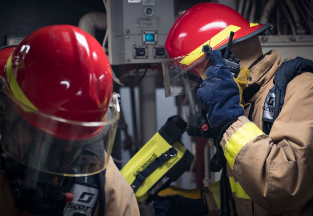 Zwei Feuerwehrleute in Schutzausrüstung arbeiten an einem Hydranten während einer übung, mit verschiedenen Maschinen und Kabeln im Hintergrund.
