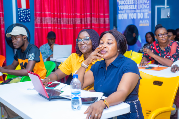 Eine Gruppe von Menschen sitzt um einen Tisch mit Laptops, Flaschen, Mobiltelefonen, Papieren, Stiften und anderen Gegenständen. Im Hintergrund ist eine Wand mit einem Vorhang und einem Banner mit Text sowie Fahnen zu sehen bei der Maya-Führungsveranstaltung 2019.