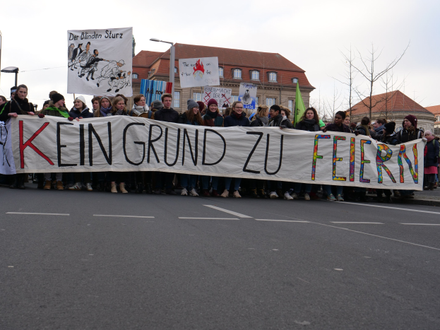 Demonstranten mit einem Transparent "Kein Grund zu Feiern" gegen deutsche Sparmaßnahmen, mit Straßeninfrastruktur und Gebäuden im Hintergrund unter einem klaren Himmel.