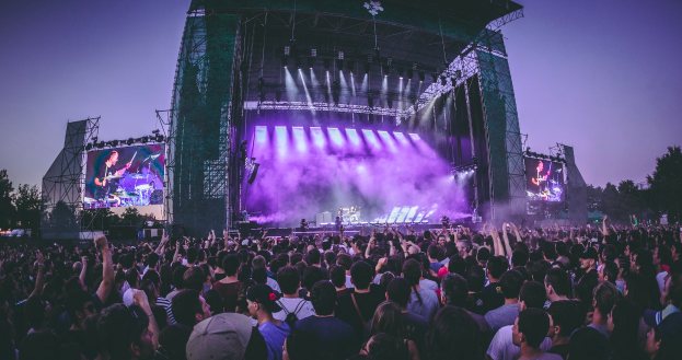 Eine große Menschenmenge versammelte sich auf einem Musikfestival, mit einer Bühne in der Mitte, auf der Musiker Instrumente spielten, beleuchtet von Scheinwerfern und umgeben von Bildschirmen, mit einem Gebäude, Bäumen und einem klaren blauen Himmel im Hintergrund.