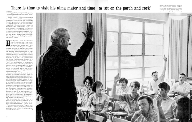 Eine Person mit Brille steht vor einer Gruppe sitzender Personen, mit einer Vorhangwand und Fenstern im Hintergrund.