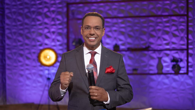 Stephen Colbert in Anzug, Krawatte und Brille auf einer Bühne mit Mikrofon, Wand, Ständer, Licht und diversen Gegenständen dahinter.