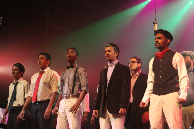 Eine Gruppe von M√§nnern auf einer B√ľhne, die ein Drama mit gr√ľnen Scheinwerfern hinter ihnen auff√ľhren.