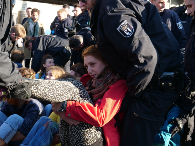 Eine Frau in dunkler Kleidung wird von einem Polizeibeamten geführt, mit einer Menge sitzender und stehender Menschen im Hintergrund, einer Hütte und einem klaren blauen Himmel.