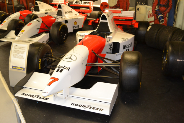 Eine Museumsausstellung von Rennwagen mit Menschen, die zuschauen, mit informativem Text auf einer Tafel, einer Seilabsperrung und einer Wand im Hintergrund.
