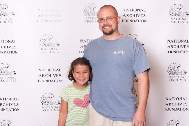 Ein Mann in einem blauen T-Shirt und cremefarbenen Hosen steht auf einem roten Teppich neben einem strahlenden kleinen Mädchen, mit einem weißen Banner mit Text und Logos im Hintergrund.