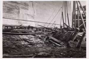 Ein Schwarz-Weiß-Foto einer Baustelle für das New Yorker Staatsgefängnis, das Arbeiter, hölzerne Gegenstände, Pfähle, Seile, eine Wand und Bäume mit einem Gebäude im Hintergrund zeigt.