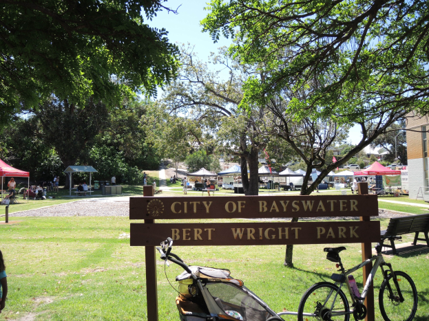 Bayswater Bert Wright Park mit einem Schild, Fahrrad, Kinderwagen, Gras, Bank, Menschen, Zelten, einem Gebäude, Straßenpfosten und einem bewölkten Himmel.