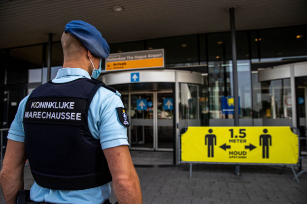 Ein Polizeibeamter in blauer Uniform und Mütze steht vor einem gläsernen Gebäude mit einem gelben Schild auf der rechten Seite und weiteren Schildern im Hintergrund.