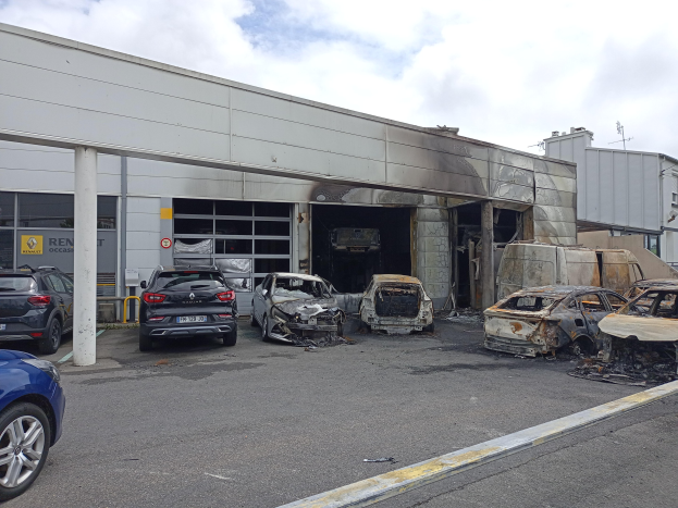 Verkohlte Autos vor einem feuerbeschädigten Gebäude mit Pfählen und Trümmern in einem bewölkten Himmel.