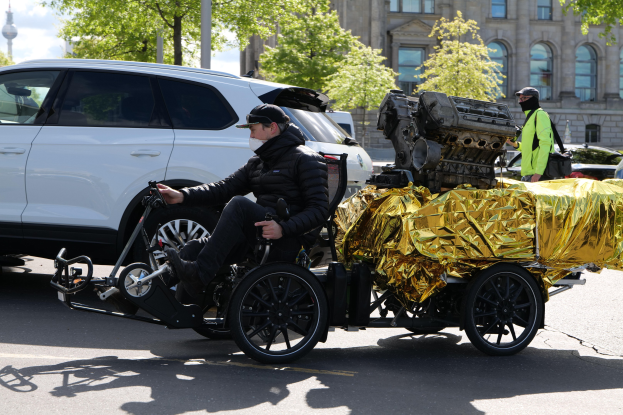 Ein Mann in einem Rollstuhl mit einem großen Motor an der Rückenlehne, umgeben von Fahrzeugen auf einer Straße mit Bäumen, Gebäuden und einem klaren blauen Himmel im Hintergrund.