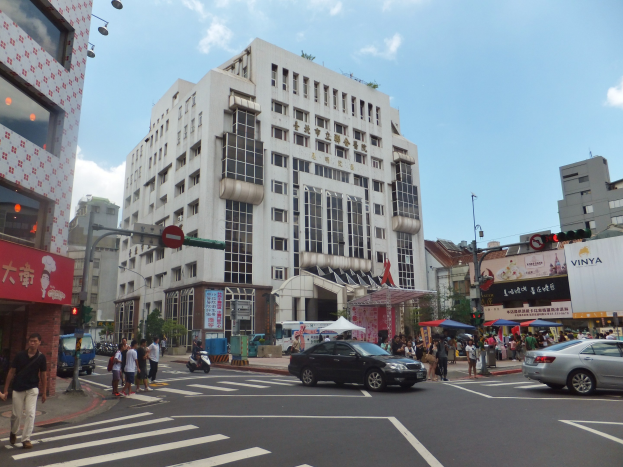 Eine belebte Straße mit Fussg"angern, fahrenden Fahrzeugen, Ampeln, hohen Geb"uden, Gehwegen, B"umen, Schirmen und einem bew"olkten Himmel.