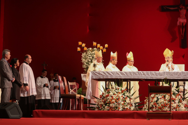 Vier Personen in einem Raum mit Kerzen auf Tischen, eine Person hält ein Mikrofon, Stühle, ein Lautsprecher, Blumen und Kreuze an der Wand im Hintergrund.