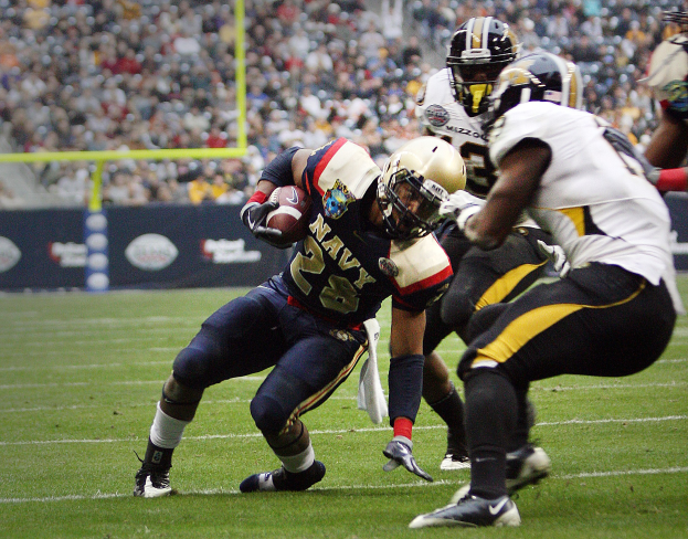 Ein Fußballspieler in Uniform rennt mit dem Ball, während ihn Gegenspieler in Sportbekleidung und Helmen verfolgen, während sie auf einem Rasenfeld spielen.