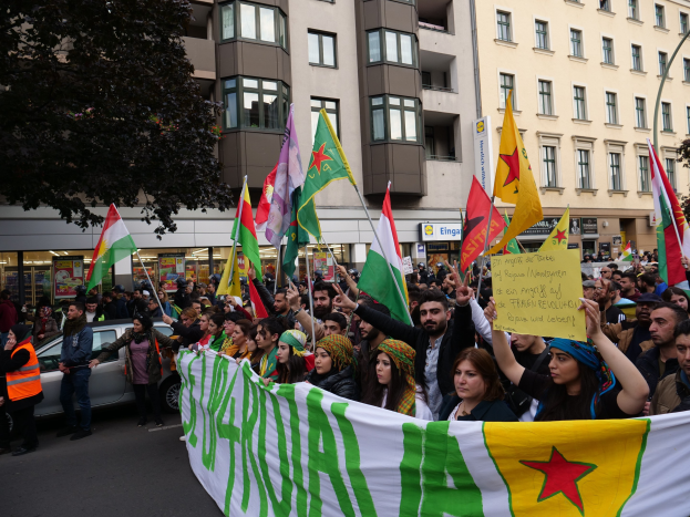 Eine große Gruppe von Menschen marschiert mit Flaggen und Schildern die Straße hinunter, mit einem geparkten Auto auf der rechten Seite und einem Baum auf der linken Seite, vor Gebäuden mit Fenstern und Namensschildern, was auf eine algerische Demonstration hinweist.