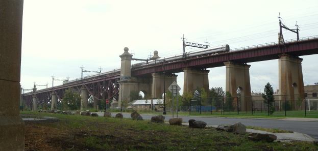 Ein Zug fährt über eine Brücke mit Säulen, Strommasten mit Drähten, Schilder, einen Zaun, Gras, Steine, eine Straße, Bäume, Gebäude und einen bewölkten Himmel im Hintergrund.
