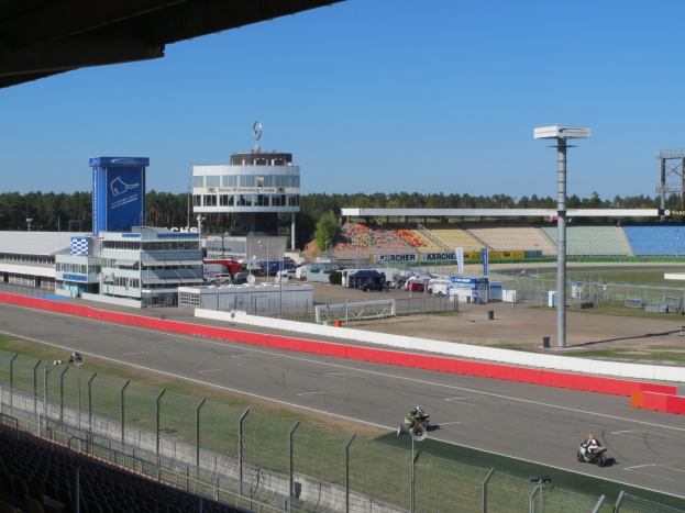 Gruppe von Motorradfahrern auf einer Rennstrecke, umgeben von einer Absperrung, Fahrzeugen, einem Gebäude, Schildern, Stadion und Bäumen unter einem bewölkten Himmel.