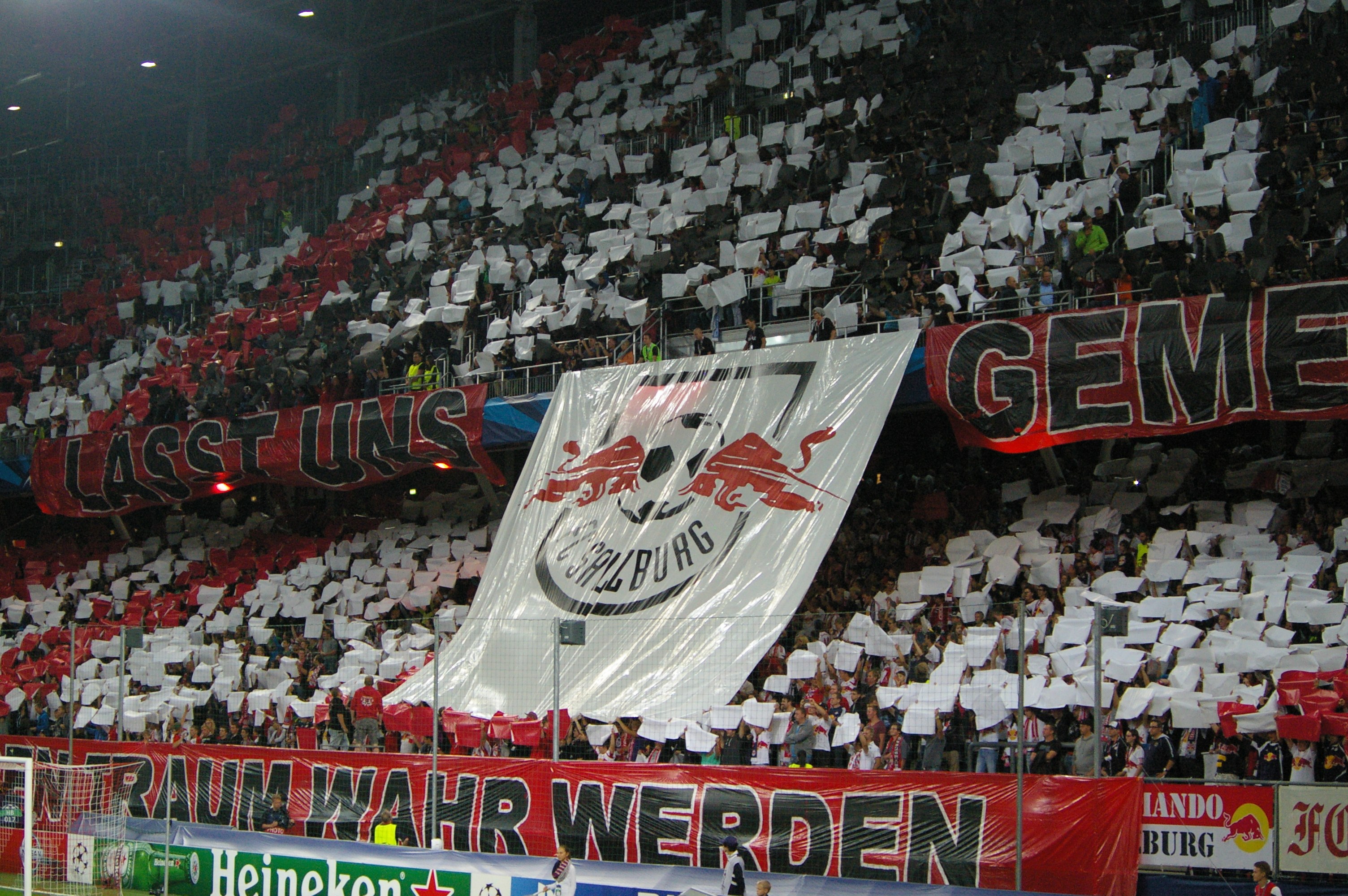 Ein volles Fußballstadion mit Sitz- und Stehplätzen, Banner, einem Torpfosten und Überdachungsbeleuchtung.