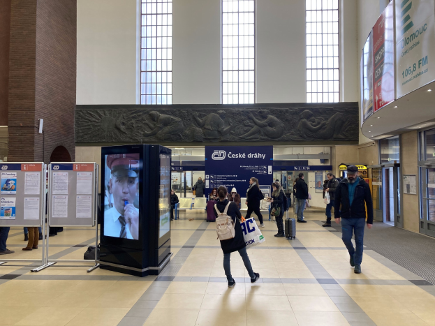 Menschen, die durch einen Bahnhof gehen mit digitalen Kiosken, Anzeigetafeln und Reliefs im Hintergrund.