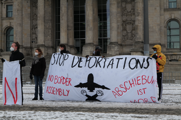 Eine Gruppe von Menschen steht im Schnee und hält ein 'Stoppt Abschiebungen'-Schild hoch und trägt Masken, mit Geländern im Hintergrund.