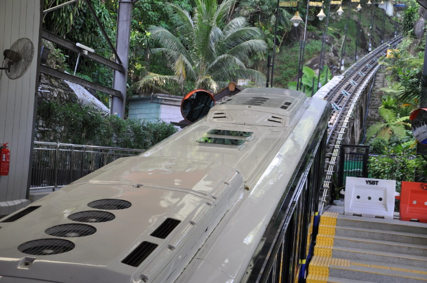 Ein Zug fährt auf Eisenbahnschienen mit Gebäuden, Masten, einem Ventilator, einem Feuerlöscher und einem Geländer auf der linken Seite, Gegenständen und einer Treppe auf der rechten Seite und Bäumen im Hintergrund.