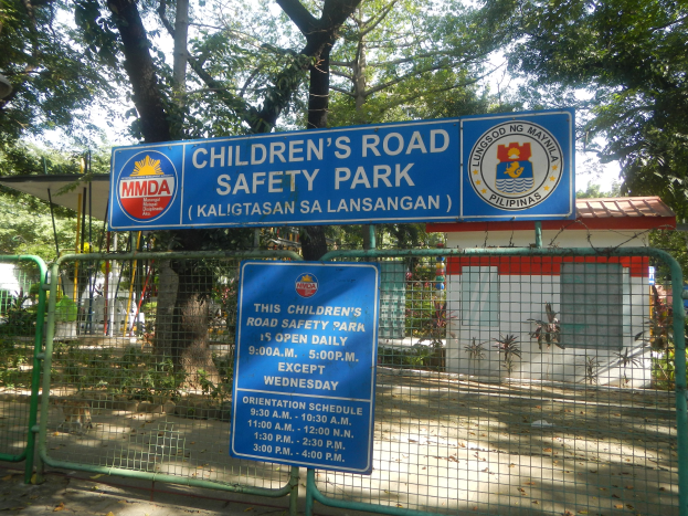 Metallzaun um einen Kinderverkehrssicherheitspark, zwei blaue Informationsschilder, ein kleines Haus mit Dach, Pflanzen, Bäume und einen bewölktem Himmel.