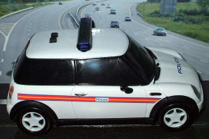 Ein weißes Polizeiauto steht im Vordergrund mit mehreren Fahrzeugen, die sich auf der Straße dahinter bewegen, umgeben von Gras und Bäumen auf beiden Seiten.