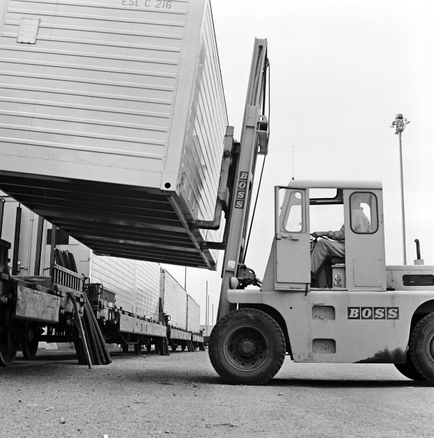 Ein Mann, der einen Stapler mit einem Container auf dem Rücken fährt, umgeben von anderen Fahrzeugen auf der Straße mit Pfählen und einem klaren Himmel im Hintergrund.