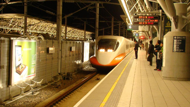 Mehrere Personen stehen auf einem Bahnsteig mit einem Zug auf den Schienen; links hat eine Wand ein Schild, rechts haben die Pfosten Schilder.