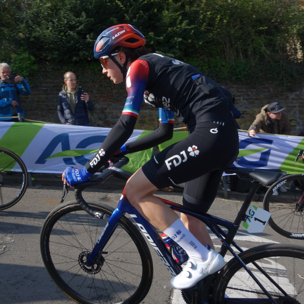 Professionelle Radrennfahrerin Katie Archibald fährt ein Fahrrad die Straße entlang, trägt einen Helm, eine Schutzbrille, Handschuhe und Schuhe, mit einer Menge Menschen und einem Banner im Hintergrund.
