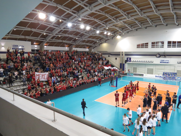 Eine Gruppe von Menschen spielt Volleyball in einer Turnhalle mit einem Netz, Zuschauern auf Stühlen mit Bannern und einer Wand mit Fenstern und Deckenbeleuchtung im Hintergrund.