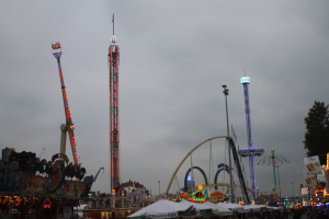 Eine Menschenmenge versammelt sich um einen nächtlichen Fasching, beleuchtet von den Lichtern der Fahrgeschäfte, Pfähle und Bäume, unter einem bewölkten Himmel.