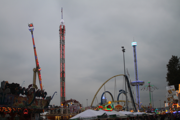 Eine Menschenmenge versammelt sich um einen nächtlichen Fasching, beleuchtet von den Lichtern der Fahrgeschäfte, Pfähle und Bäume, unter einem bewölkten Himmel.
