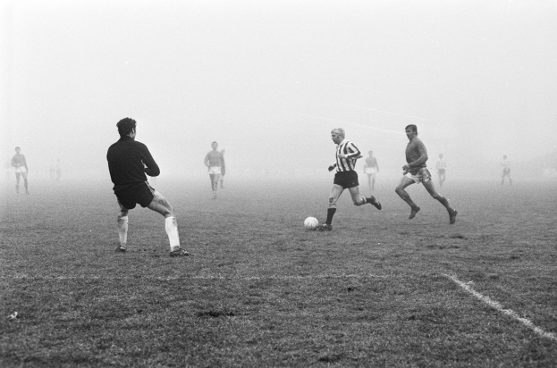 Eine Gruppe von Männern, die auf einem nebligen Feld Fußball spielen, mit einem Ball in der Mitte der Aktion, dargestellt in Schwarz-Weiß.