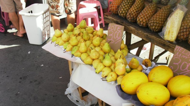 Eine Marktszene mit verschiedenen Fruächten auf einem Gestell, einigen Hockern, ein paar Menschen und zwei Preiszetteln.