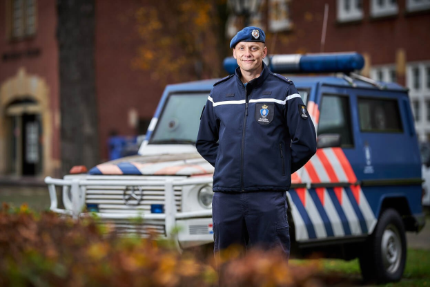 Ein Polizist in Uniform steht vor einem Polizeiwagen, trägt eine Mütze und hat einen unscharfen Hintergrund und Pflanzen unten.