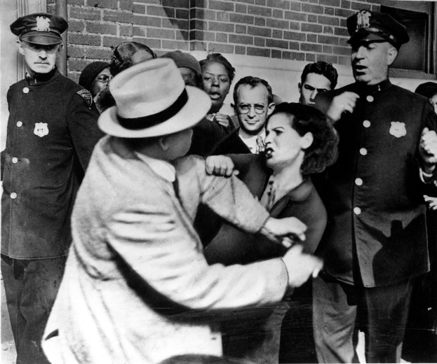 Zwei Personen kämpfen in einem Schwarz-Weiß-Bild, im Hintergrund sind mehrere andere zu sehen, einige tragen Mützen, und eine Wand im Hintergrund.